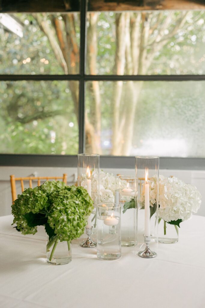 A white table with green and white hydrangeas in vases, tall glass candle holders, and white taper candles, set by a large window with trees outside—an elegant inspiration for wedding reception venues Atlanta couples will love.