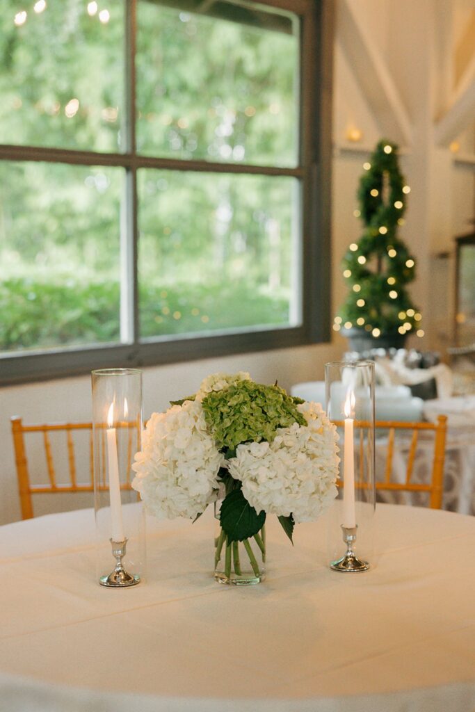 A round table with a white cloth holds a bouquet of white and green hydrangeas in a vase, flanked by two tall glass candle holders. Perfect for those seeking elegant wedding reception venues Atlanta has to offer, the scene is set against a large window and features a lit, spiral topiary in the background.