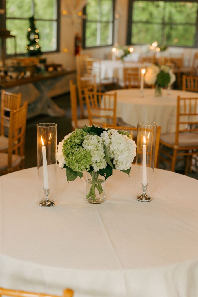 A round table with a white tablecloth, a vase of white and green hydrangeas, and two lit candles in glass holders creates an elegant setting often seen at top wedding reception venues Atlanta offers. Wooden chairs and more decorated tables are visible in the background.
