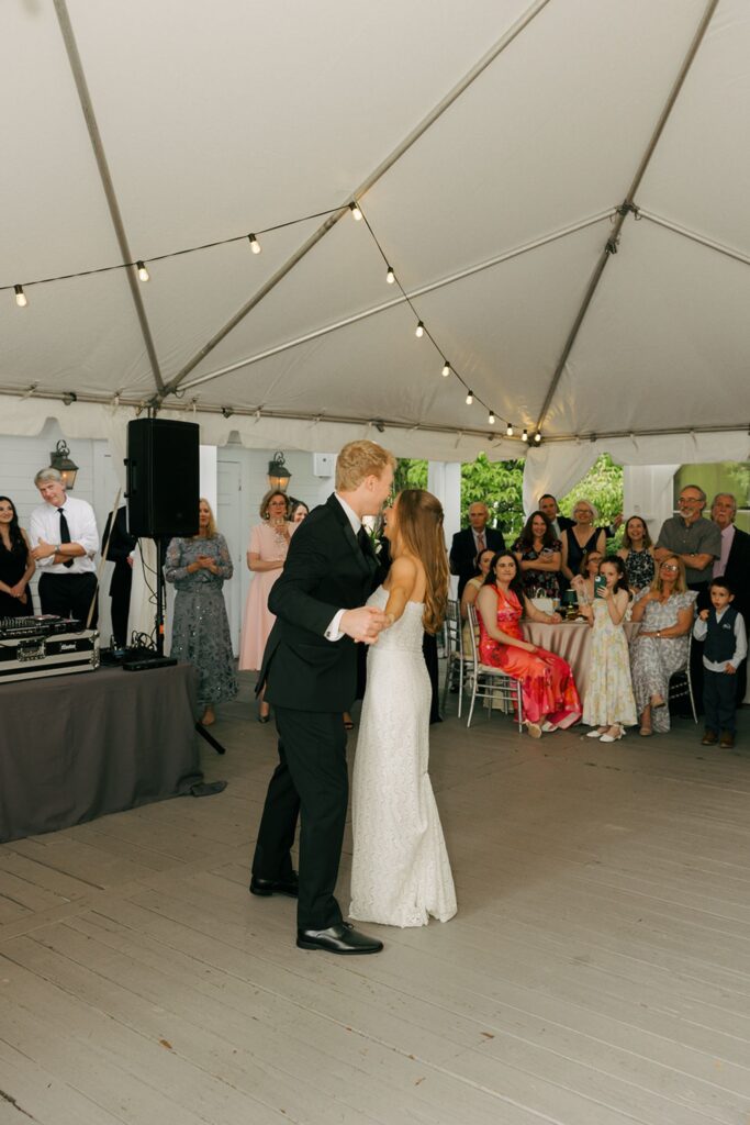 A bride and groom share their first dance under a white tent at one of the elegant wedding reception venues Atlanta has to offer, surrounded by seated and standing guests who watch and smile. String lights hang above, and a DJ stands by his setup in the background.