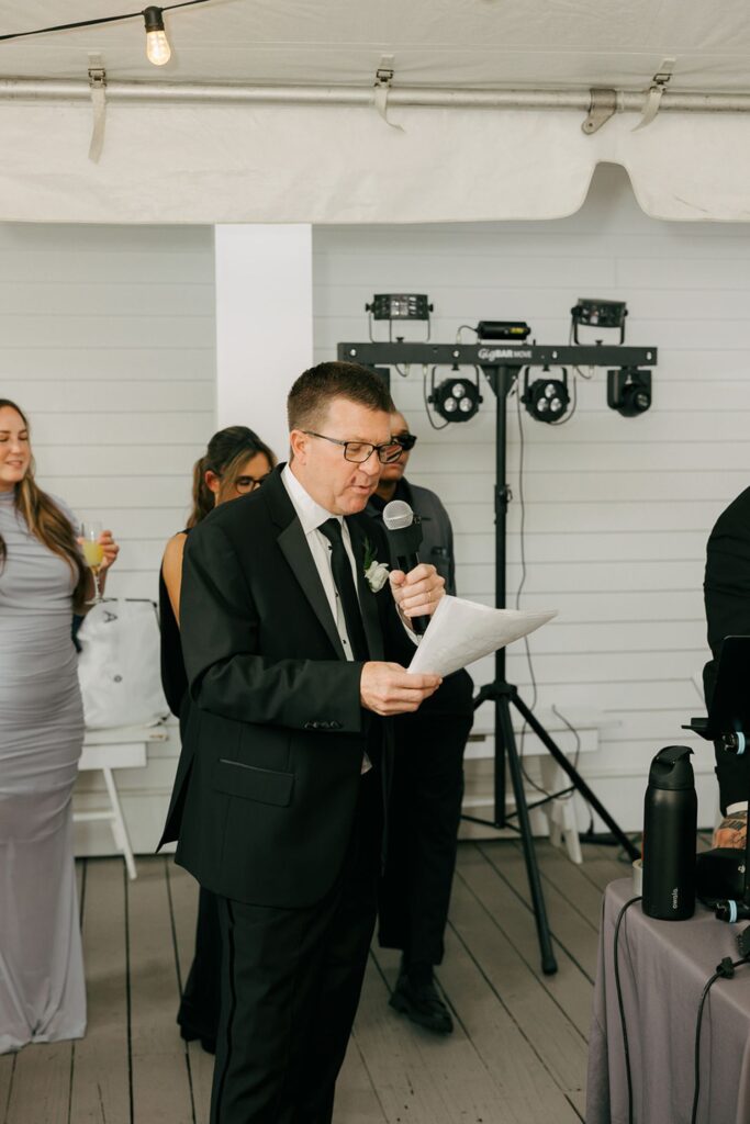 A man in a black suit and glasses reads from a paper while speaking into a microphone at an indoor event, possibly one of the elegant wedding reception venues Atlanta is known for. People stand in the background, and event equipment is visible behind him.