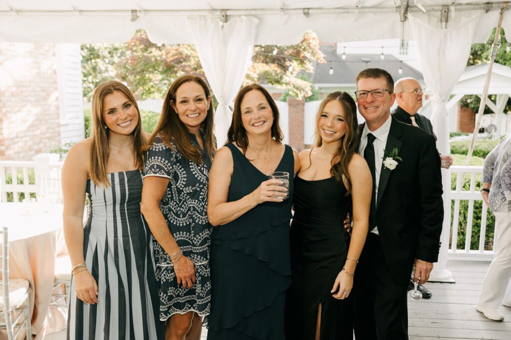Five people dressed in formal attire smile together under a white tent at an outdoor event, with greenery and buildings visible in the background—an elegant setting reminiscent of top wedding reception venues Atlanta has to offer.
