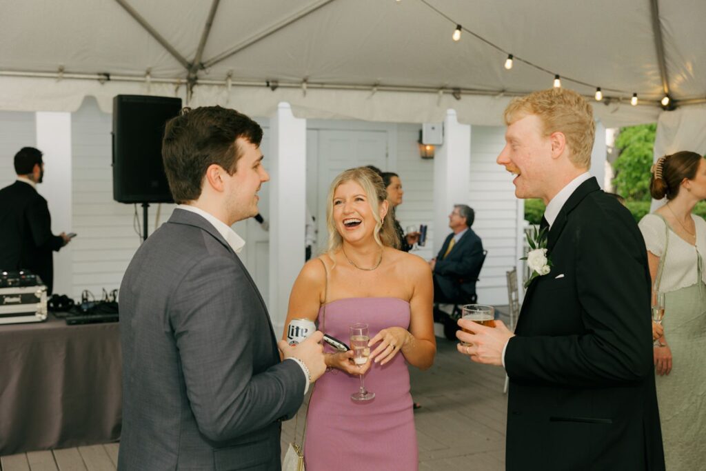 Three people dressed formally stand under a tent, smiling and talking at a party. One woman in a lavender dress holds a drink, while two men in suits chat with her, all appearing happy and relaxed—capturing the joyful atmosphere often found at wedding reception venues Atlanta couples adore.