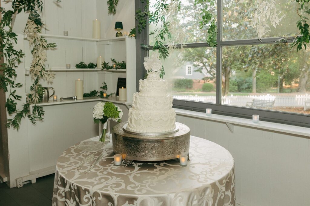 A white, ornate wedding cake sits on a silver stand atop a round, patterned tablecloth. Greenery, candles, and a vase with white flowers decorate the room near a large window—creating an enchanting atmosphere reminiscent of the finest wedding reception venues Atlanta has to offer.