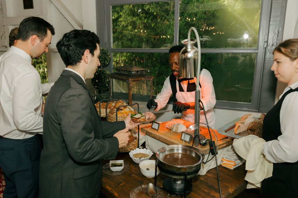 A chef slices meat at a carving station while two men in suits wait in line—typical of the elegant atmosphere found at many wedding reception venues Atlanta offers. A woman in uniform stands by, and baskets of bread are displayed on the table near a window with greenery outside.