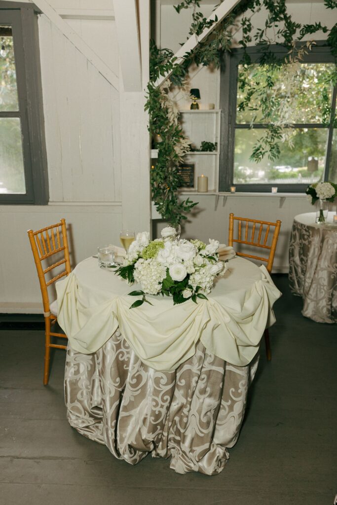 A round table with a cream and beige patterned tablecloth, a floral centerpiece of white flowers, wooden chairs, and greenery decorates a cozy, softly lit indoor space with large windows—capturing the elegant charm found in many wedding reception venues Atlanta has to offer.
