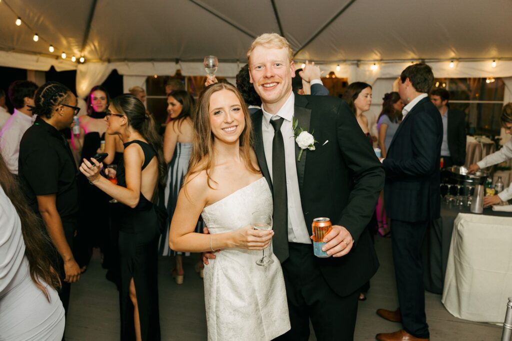A smiling couple poses at a party; the woman wears a white dress and holds a wine glass, while the man in a suit holds a can. Other guests mingle in the background under a tent with string lights, creating an atmosphere reminiscent of elegant wedding reception venues Atlanta is known for.