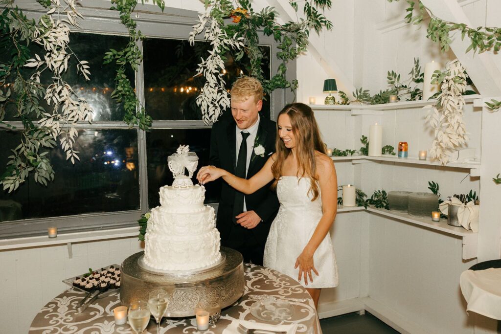 A bride and groom, smiling, cut a tiered white wedding cake together in a beautifully decorated room with leafy greenery and candles—one of the most charming moments often seen at wedding reception venues Atlanta couples love.