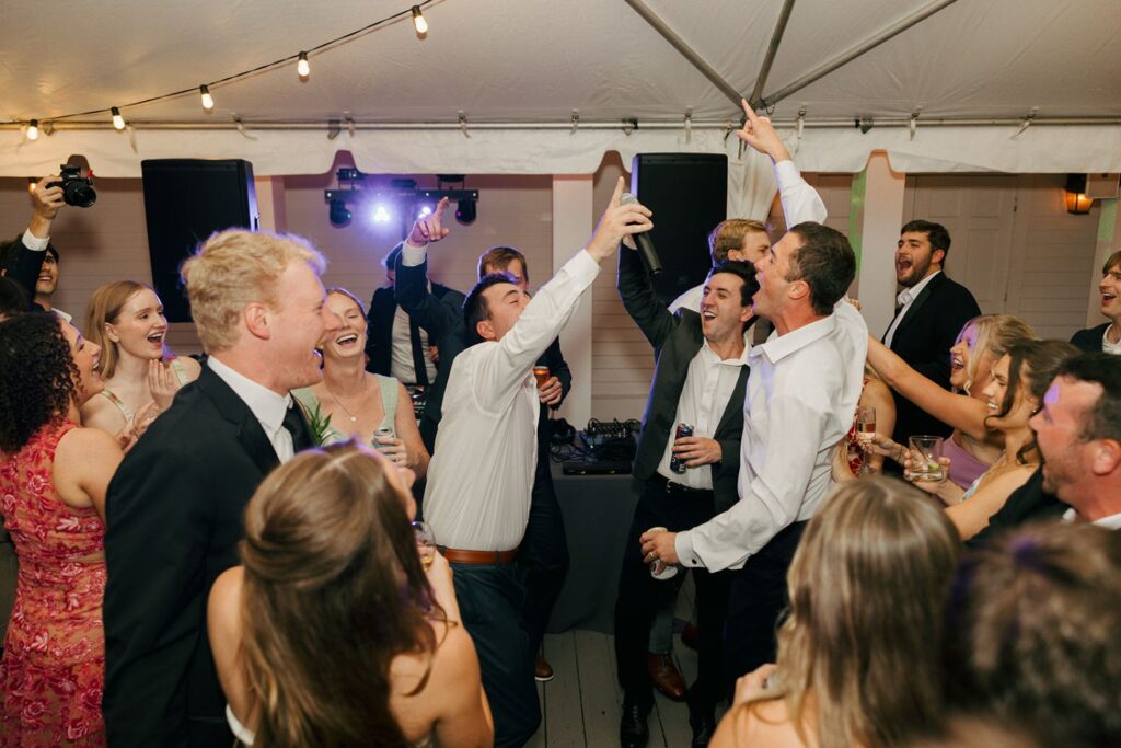 A lively group of people dressed formally sing and celebrate together under a tent with string lights, some raising glasses and phones, enjoying a festive atmosphere at one of the most charming wedding reception venues Atlanta has to offer.