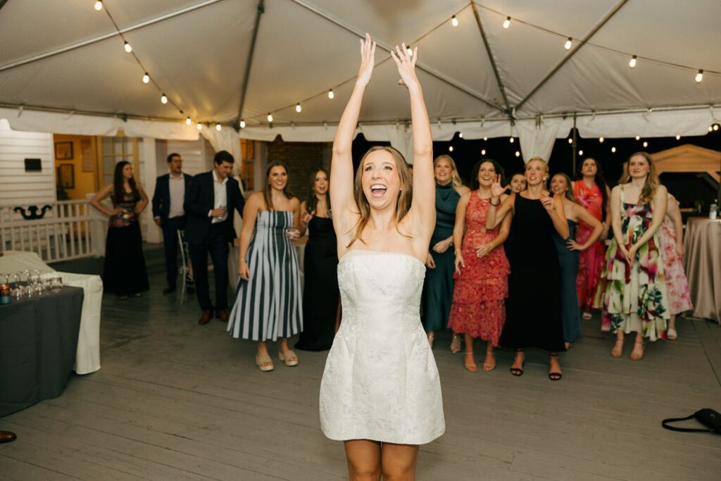 A woman in a white dress raises her arms to toss a bouquet, with a group of women behind her, ready to catch it, at a festive event under a string-lit tent—one of the many lively moments that make wedding reception venues Atlanta such memorable places to celebrate.