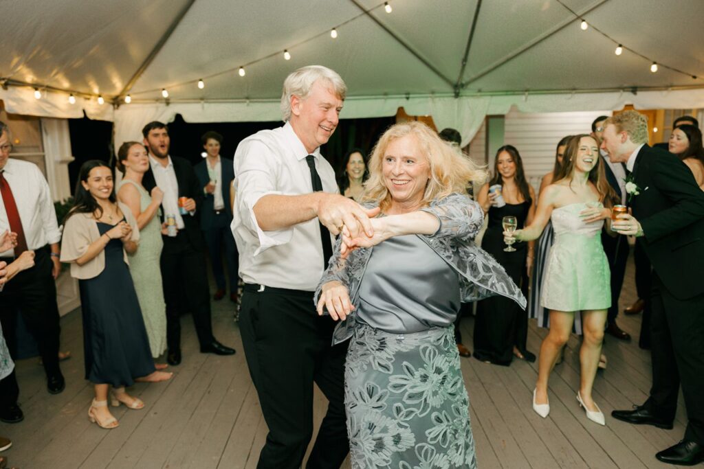 A smiling man and woman dance together under a tent at a festive event, surrounded by guests watching and enjoying themselves. String lights hang above, creating a warm and lively atmosphere reminiscent of the best wedding reception venues Atlanta has to offer.