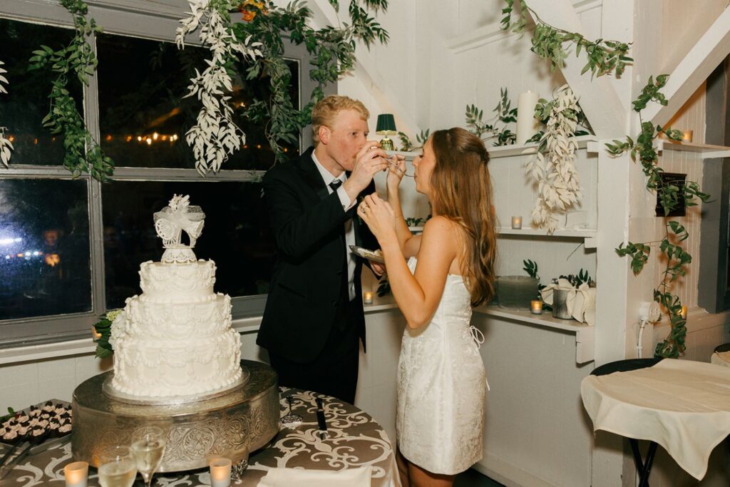 A couple in formal attire feeds each other cake at a wedding reception, standing beside a decorated tiered cake in a room adorned with greenery and candles—an elegant setting that reflects the charm of top wedding reception venues Atlanta has to offer.