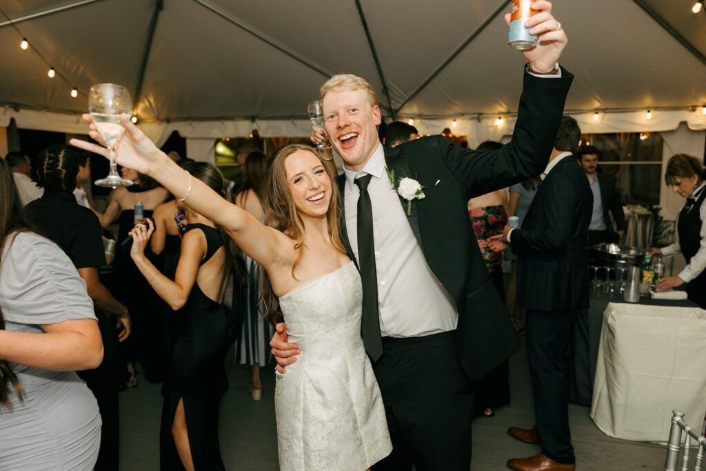 A joyful couple, dressed formally, smile and pose with raised drinks at a lively indoor celebration, surrounded by other guests under a white tent with string lights—an enchanting scene reminiscent of the best wedding reception venues Atlanta has to offer.