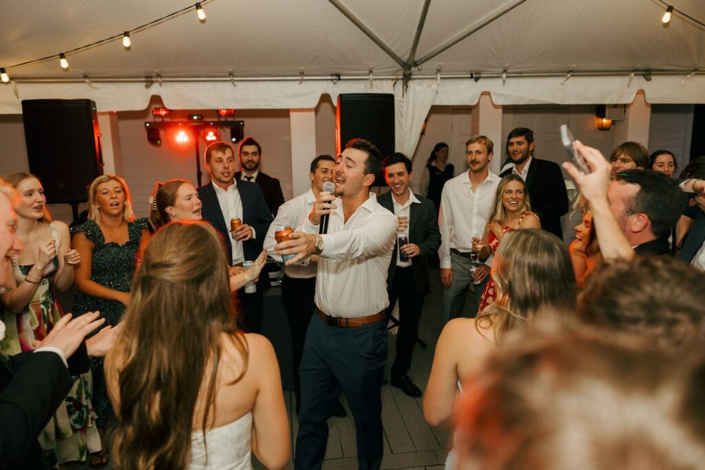 A man sings into a microphone while holding a drink, surrounded by a group of people cheering and smiling at a lively event under a white tent with string lights—capturing the joyful spirit often found at wedding reception venues in Atlanta.