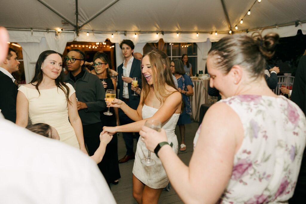 A group of people dressed formally dance and socialize under string lights at an indoor event, showcasing the elegance often found in top wedding reception venues Atlanta has to offer. A woman in a white dress smiles and holds a drink while dancing with others around her.