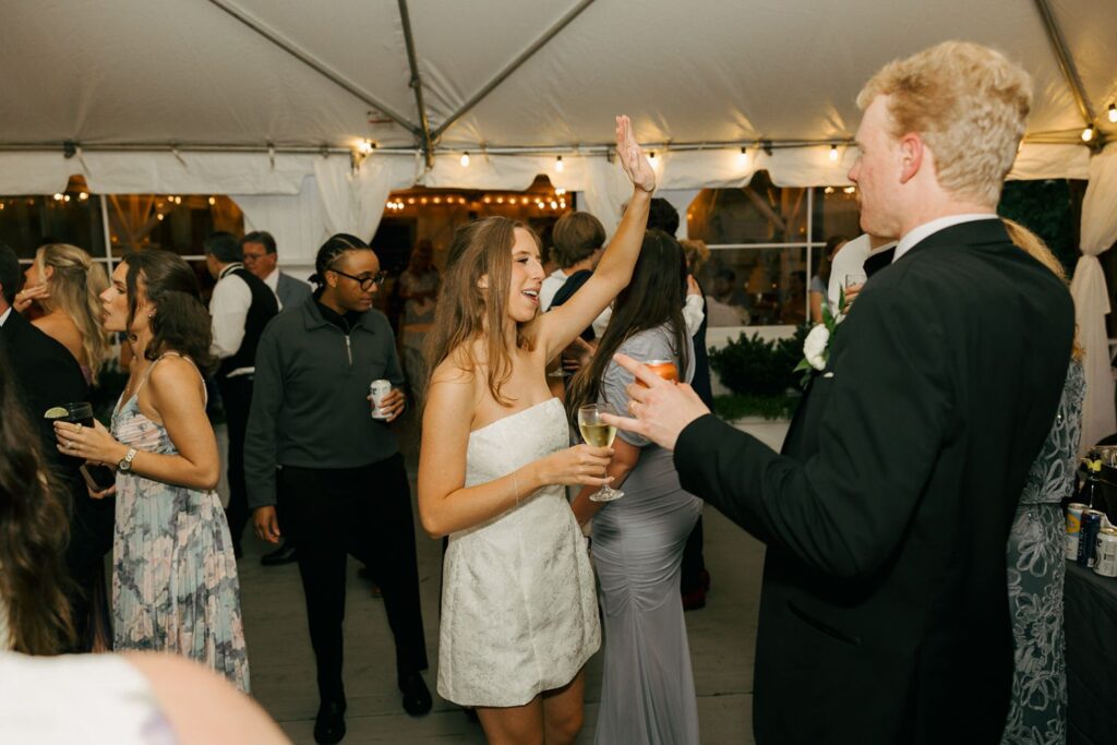 A woman in a white dress high-fives a man in a suit at a lively indoor event, perfectly capturing the joyful spirit found at top wedding reception venues Atlanta has to offer, while other guests mingle and dance under a white tent adorned with glowing string lights.