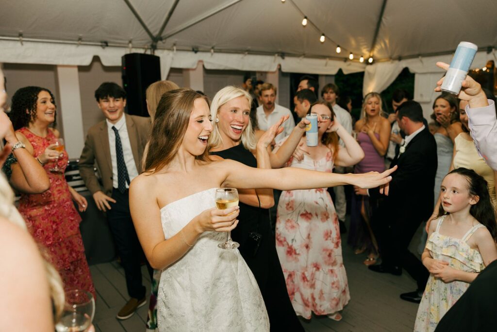 A group of people dance and celebrate at a lively wedding reception under a tent at one of the beautiful wedding reception venues Atlanta has to offer. A woman in a white dress smiles, holding a drink, while others join in and enjoy the festive atmosphere.