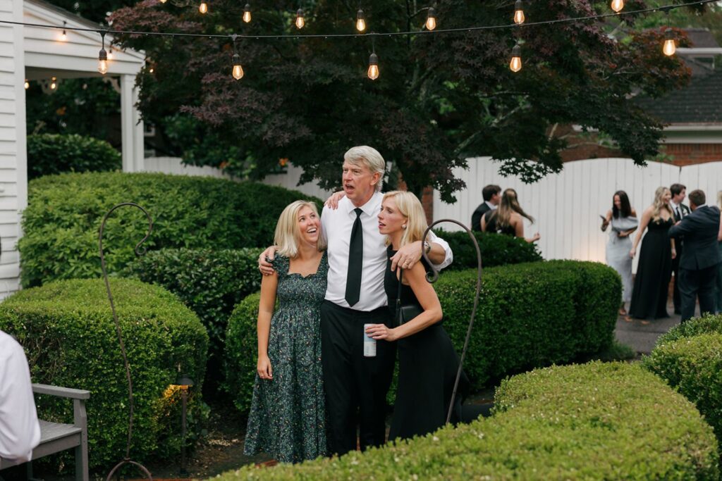 Three people dressed formally stand together outside among hedges, with string lights above—a scene reminiscent of elegant wedding reception venues Atlanta is known for. The man in the center has his arms around the two women, all smiling, as other guests mingle in the background.