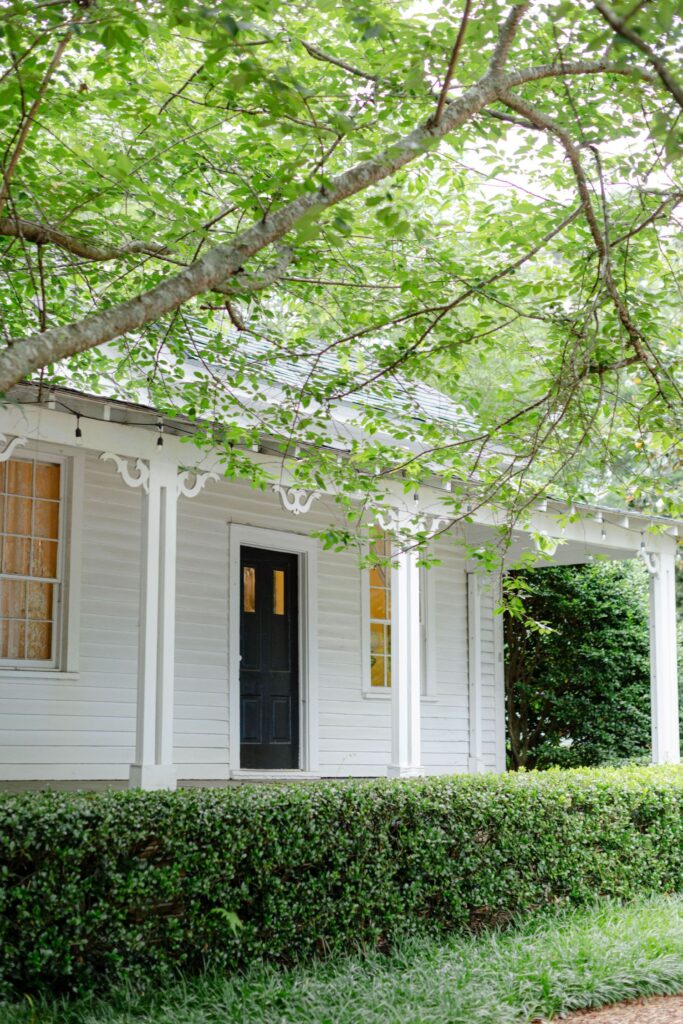 A charming white house with a covered porch, black front door, glowing windows, and lush green trees—perfect for those seeking unique rehearsal dinner venues Atlanta offers.