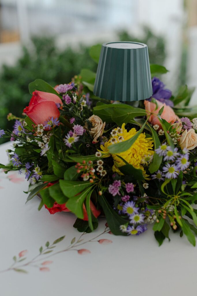 A green lamp sits in the center of a colorful floral arrangement featuring red, yellow, pink, and purple flowers—creating a charming look perfect for rehearsal dinner venues Atlanta on a white table with a subtle floral pattern.