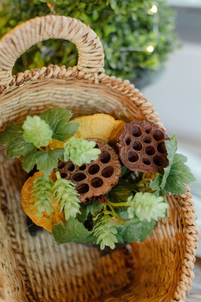 A woven basket contains green leaves, dried lotus pods, and yellowish foliage—perfect inspiration for rehearsal dinner venues Atlanta with botanical décor, with a green plant blurred in the background.