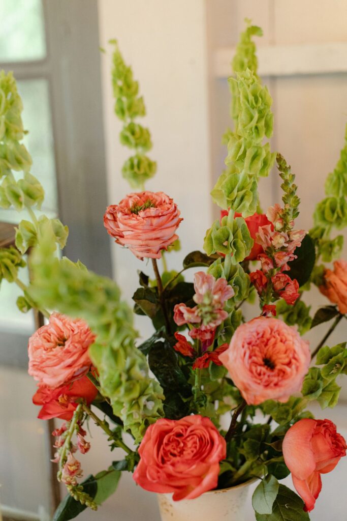 A bouquet of pink and peach roses mixed with tall green bells of Ireland flowers in a white vase, perfect for brightening up rehearsal dinner venues Atlanta, set indoors near a window.