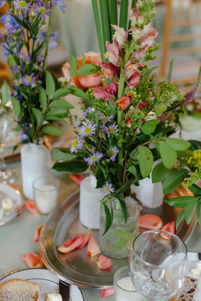 A close-up of a table setting at one of the top rehearsal dinner venues Atlanta, featuring a vase of pink, purple, and green flowers, rose petals, plates, glasses, and small candles on a silver tray.