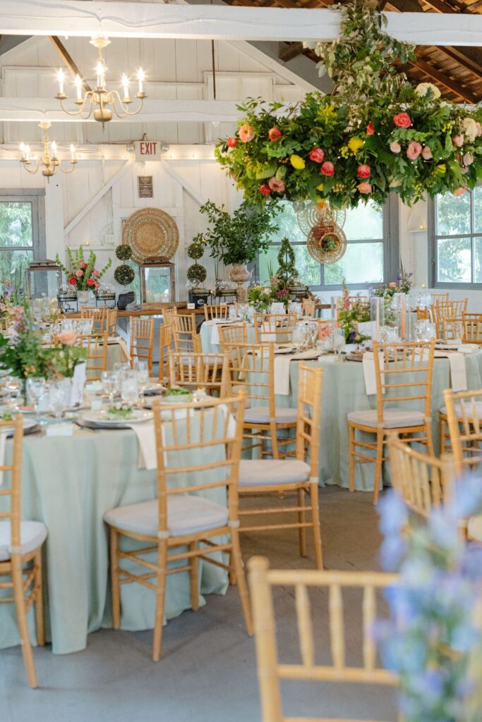 Elegant banquet hall with round tables draped in pale green cloth, wooden chairs, and chandeliers—one of the finest rehearsal dinner venues Atlanta offers, with floral centerpieces and greenery brightening the rustic space.