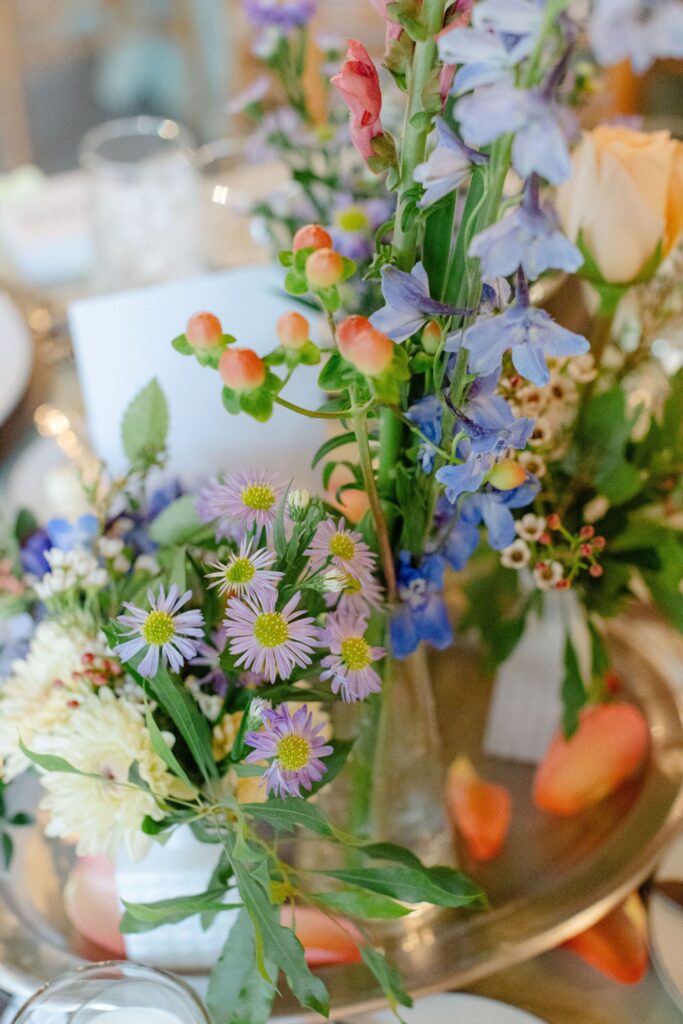 A table centerpiece with assorted fresh flowers, perfect for rehearsal dinner venues Atlanta, featuring purple asters, blue delphiniums, orange berries in glass vases, and scattered rose petals with blurred background elements.
