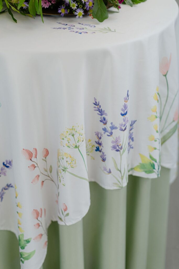 A white tablecloth with a floral print featuring pastel-colored flowers and green leaves drapes over a pale green table skirt—an elegant touch for rehearsal dinner venues Atlanta. A bouquet with purple flowers tops the table.