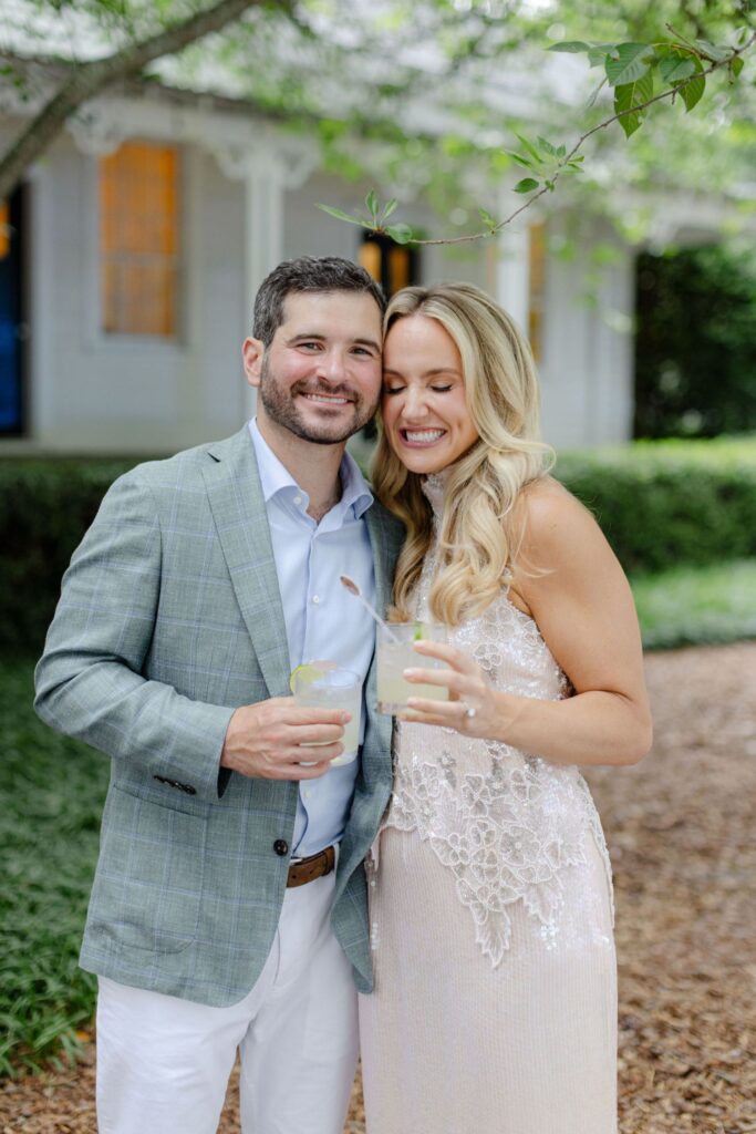 A man in a gray suit and a woman in a light pink dress smile and hold drinks while standing outside near greenery at one of the elegant rehearsal dinner venues Atlanta has to offer.