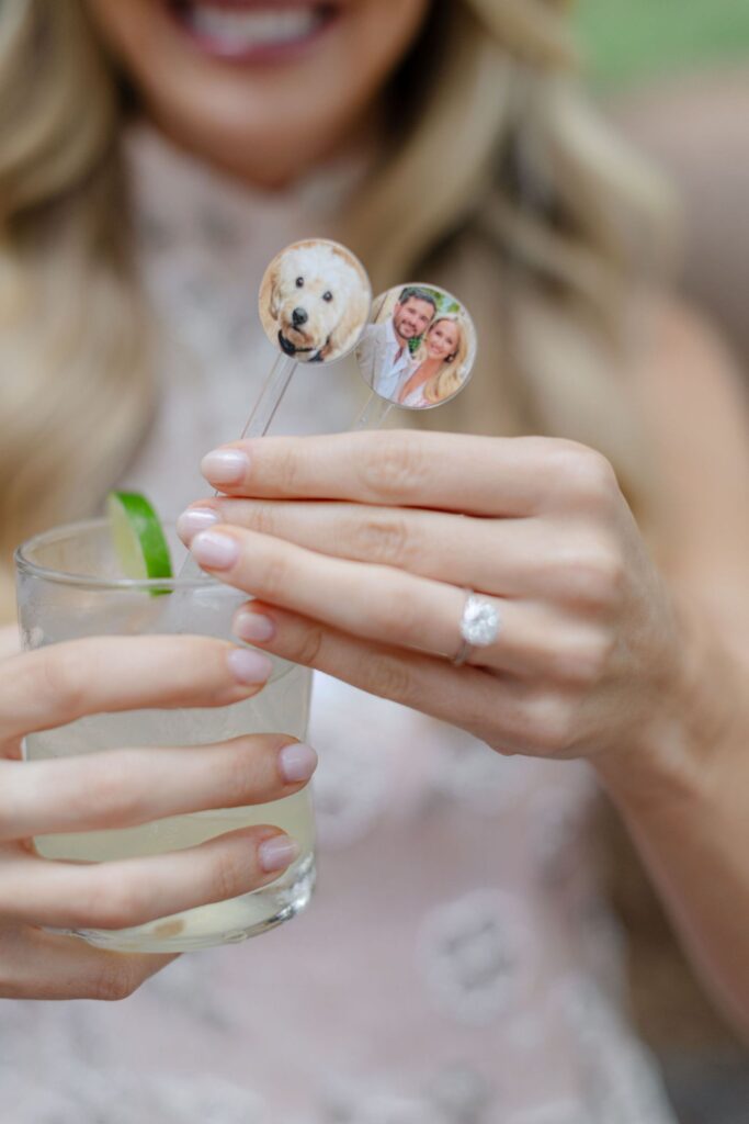 A woman with manicured nails and an engagement ring holds a drink at one of the top rehearsal dinner venues Atlanta, with two cocktail stirrers—one featuring a dog’s face and another with a smiling couple.