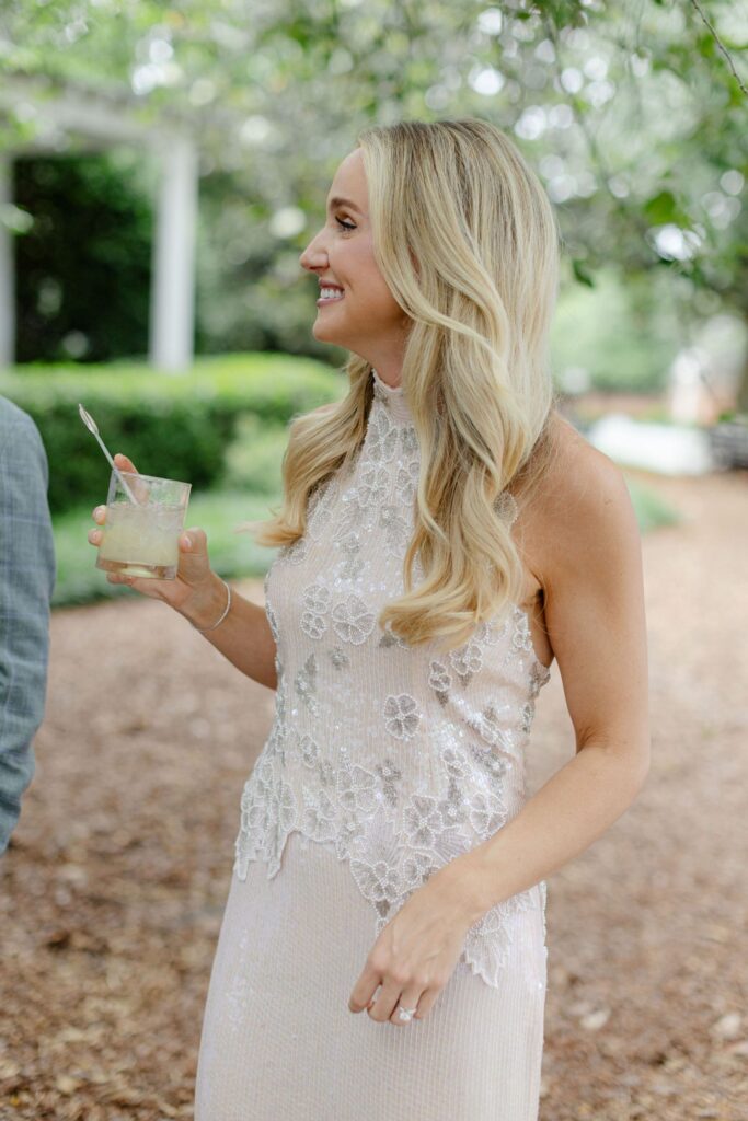 A woman with long blonde hair in a sleeveless white lace dress smiles while holding a drink outdoors at one of the elegant rehearsal dinner venues Atlanta has to offer, surrounded by greenery and a blurred background.