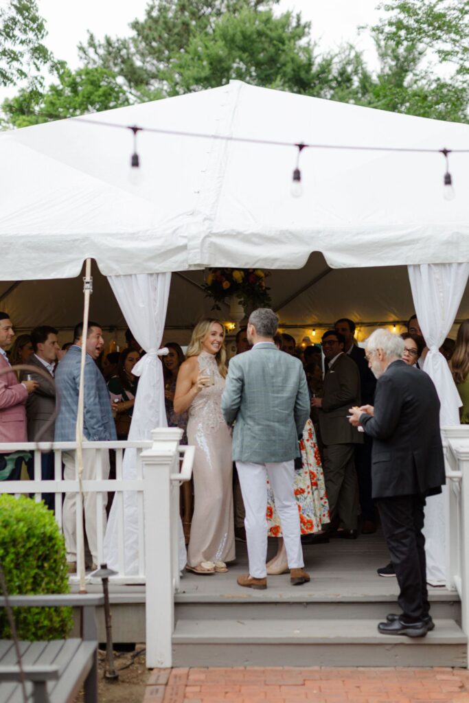 A group of people in formal attire socialize under a white tent at an outdoor event. String lights, greenery, and a wooden deck create a festive vibe—perfect for rehearsal dinner venues Atlanta offers.