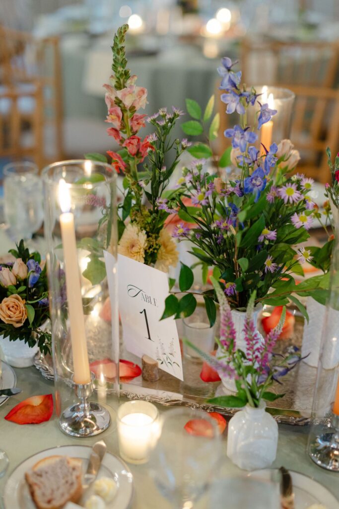 A decorated table with colorful flower arrangements, candles, scattered rose petals, a bread plate, and a card displaying Table 1—perfect for romantic rehearsal dinner venues Atlanta offers.