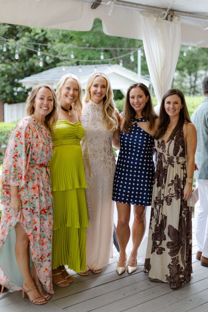Five women pose together under a tent at an outdoor event, dressed in colorful and patterned dresses, smiling at the camera—perfect inspiration for rehearsal dinner venues Atlanta with string lights and greenery in the background.