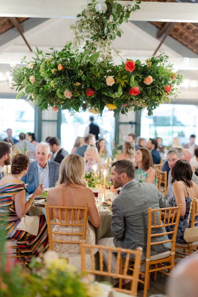 A group of people sit at decorated tables in a bright, elegant venue—one of the top rehearsal dinner venues Atlanta offers—with a large floral arrangement above as they enjoy a formal meal together.