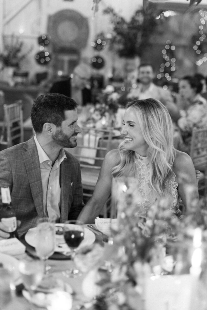 A smiling man and woman sit close together at a decorated table during a festive event, possibly exploring rehearsal dinner venues Atlanta, with drinks and food in front of them and blurred guests in the background.