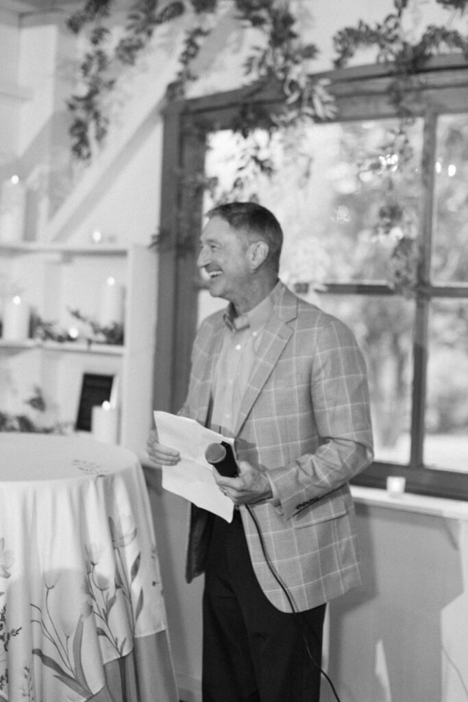 A man in a plaid blazer smiles while holding a microphone and paper, standing by a table with a floral tablecloth—capturing the joy often found at rehearsal dinner venues Atlanta offers. Greenery adorns the background.
