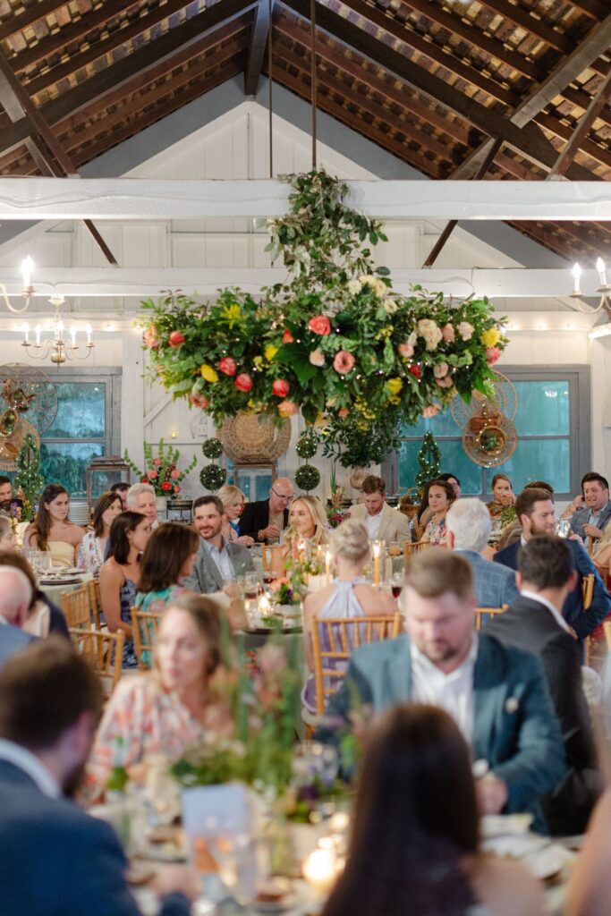 Wedding reception in a rustic hall with guests seated at decorated tables, beneath a large floral chandelier—a stunning example of what rehearsal dinner venues Atlanta can offer, with warm lighting and lush greenery for an elegant, festive atmosphere.