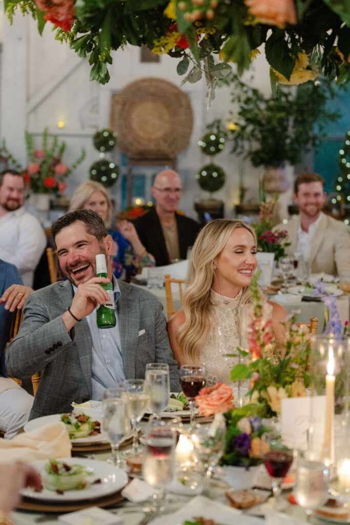 A man in a suit laughs and holds a beer, sitting beside a smiling woman at a decorated dinner table during a festive indoor event at one of the top rehearsal dinner venues Atlanta has to offer.