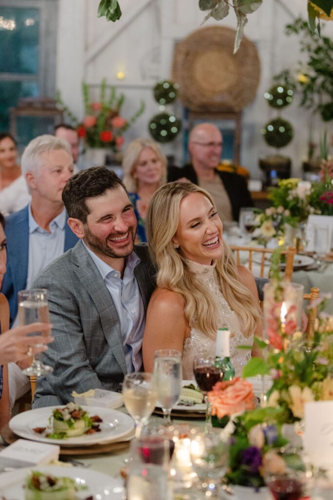 A couple sits closely together at a decorated dinner table, smiling and laughing among other guests at a festive indoor event, possibly one of the top rehearsal dinner venues Atlanta has to offer.