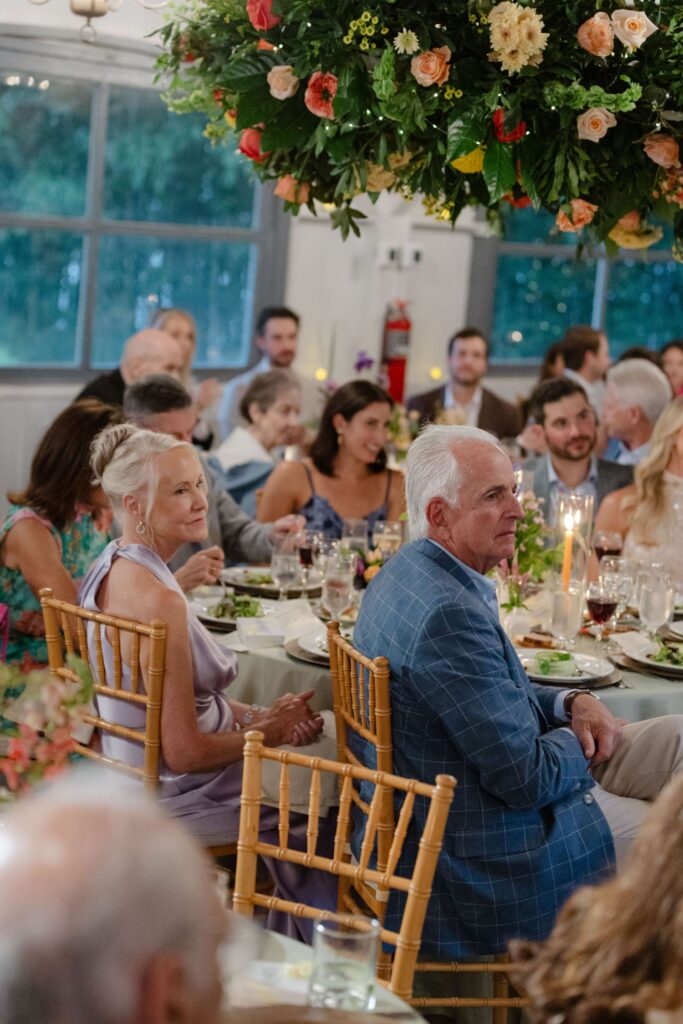 Guests seated at round tables during a formal event, listening attentively. Elegant floral decorations hang above at one of the top rehearsal dinner venues Atlanta offers, with candles and plates of salad visible on the tables.