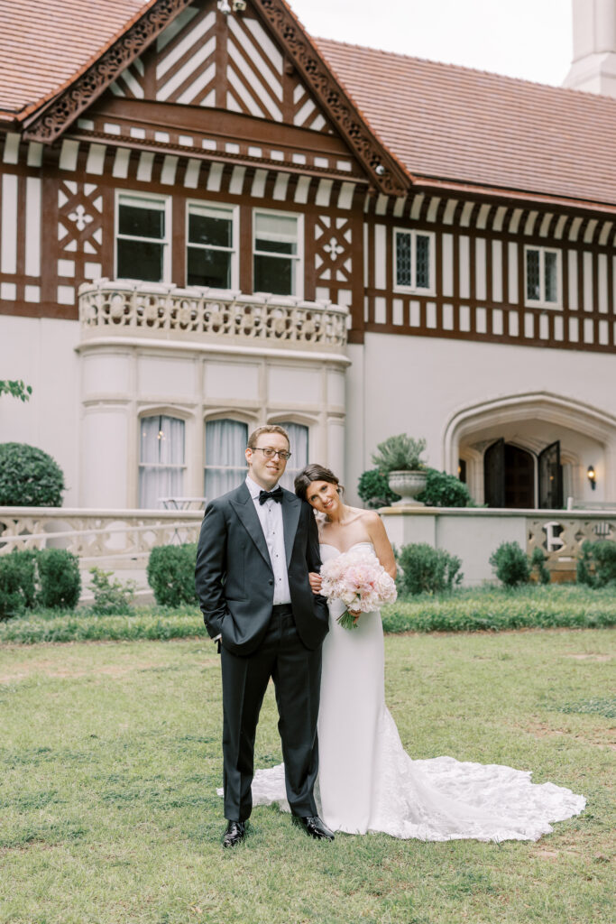 A bride in a strapless white gown leans her head on a groom in a tuxedo; they stand smiling on grass in front of a large, elegant Tudor-style building, celebrating with kosher wedding catering Atlanta.