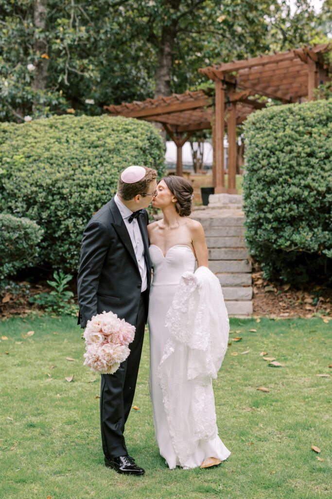 A bride and groom kiss outdoors, surrounded by greenery after their kosher wedding catering Atlanta celebration. She holds her dress and bouquet; he wears a tuxedo and kippah near steps and a wooden arbor.