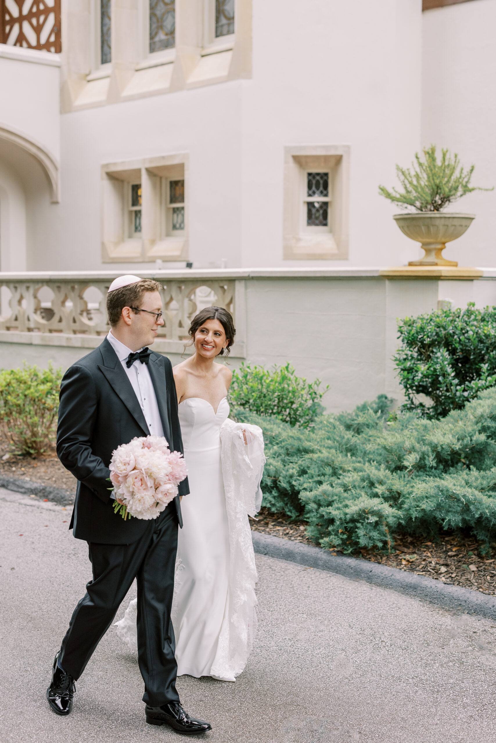 A bride in a white gown smiles at a groom in a black tuxedo holding pink flowers as they walk outdoors near a building with arched windows and greenery, celebrating with kosher wedding catering Atlanta.
