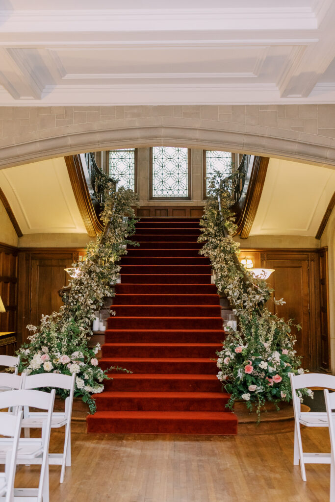 A grand staircase with a red carpet is decorated with lush green and white floral arrangements, perfect for a kosher wedding catering Atlanta event, leading up to stained glass windows and white chairs on polished wooden floors.