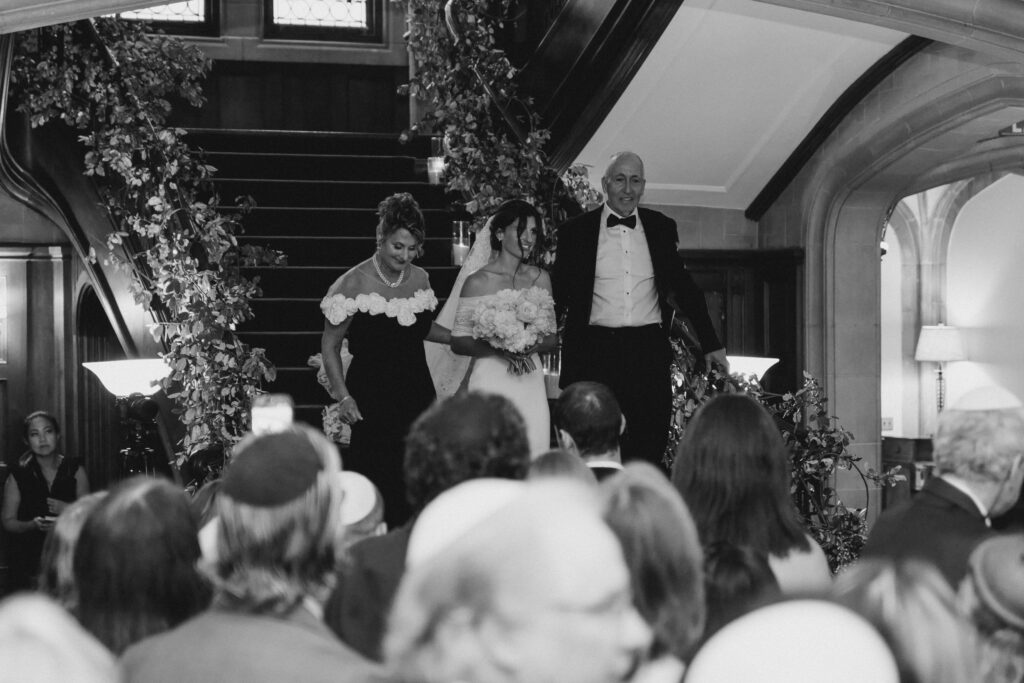 A bride in a white dress walks down the stairs with her parents, surrounded by wedding guests and elegant décor at a venue offering top-tier kosher wedding catering in Atlanta.