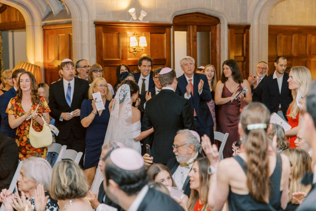 A bride and groom walk down the aisle as smiling guests stand, clap, and take photos in an elegant hall with wood-paneled walls—perfect for a kosher wedding catering Atlanta celebration.