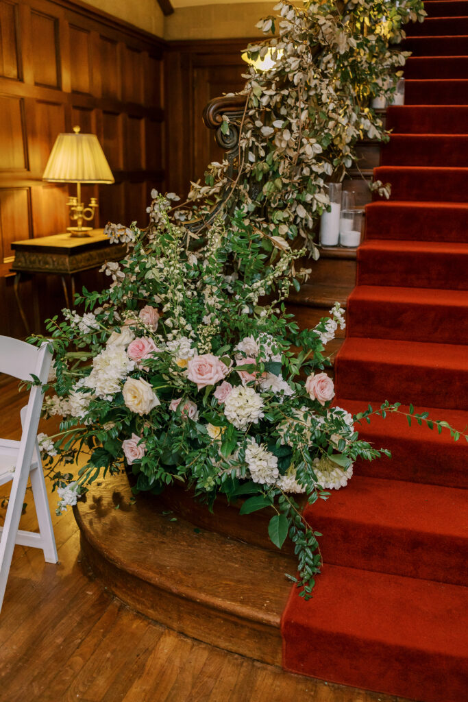 A red-carpeted staircase with ornate banister is adorned with a lush floral arrangement, perfect for a kosher wedding catering Atlanta event, in a wood-paneled room with elegant lamp and white chair nearby.